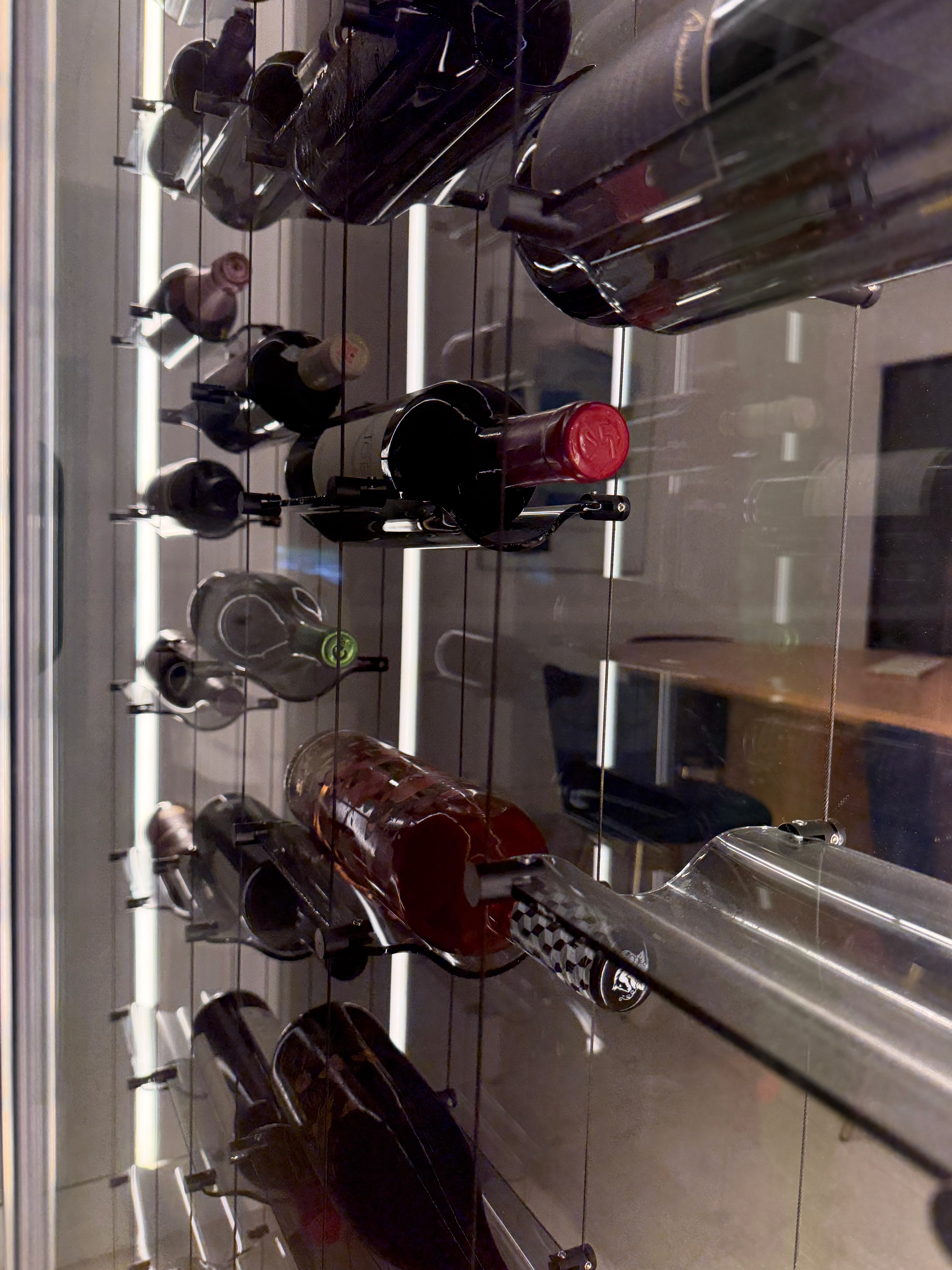 Angled interior view of floating wine bottles mounted on transparent glass supports suspended from vertical cables with LED lighting behind the installation