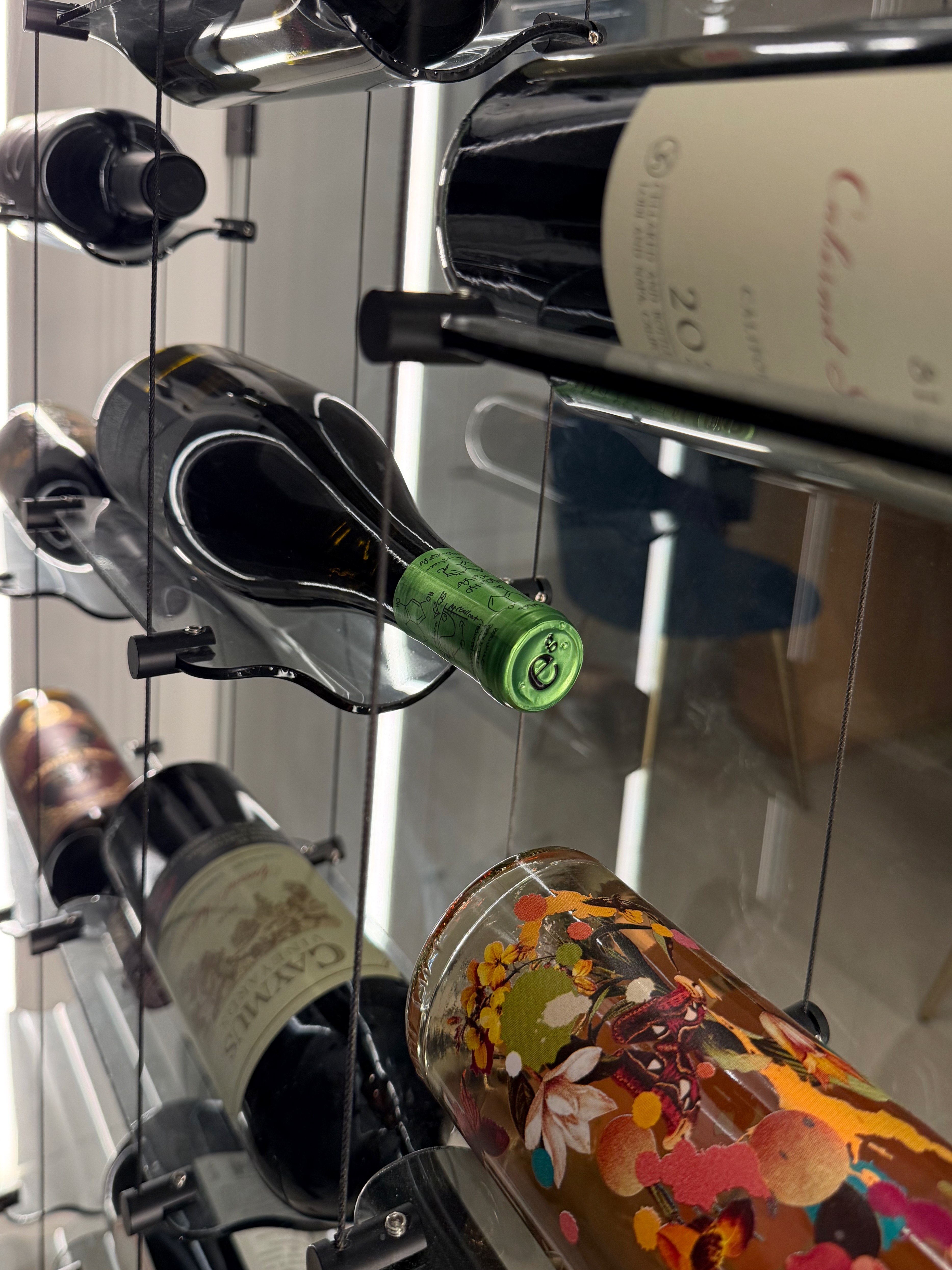 Modern dining room scene looking toward a glass wine gallery installation framed with LED lighting and floating wine bottles, with a wooden dining table and orchid in the foreground
